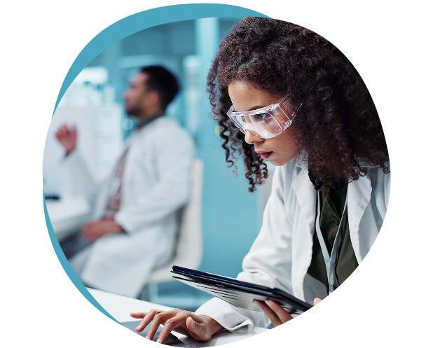 Laboratory researcher in safety glasses reviewing notes on a tablet, with a colleague in the background